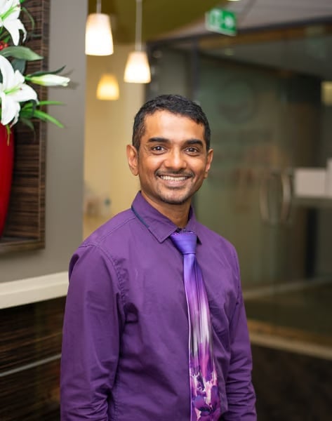 Man in purple shirt and tie smiling at camera in modern office with hanging lights and white flowers