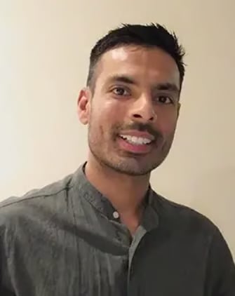 Smiling man with dark hair and facial hair wearing a gray button-up shirt against a beige wall