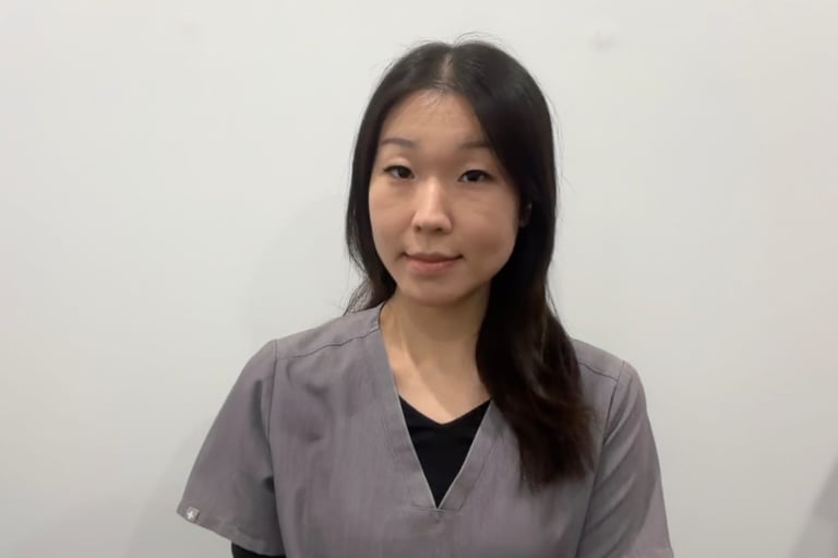 Portrait of a young woman with long dark hair wearing a gray button-up shirt over a black shirt, smiling at the camera against a white background