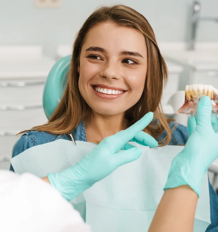 Young woman smiling in dental chair wearing turquoise gloves and showing teeth model to camera